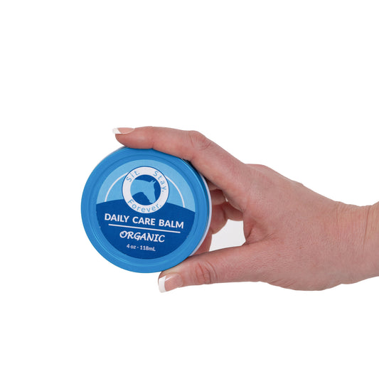 A hand holding a blue container labeled Sit. Stay. Forever. DAILY CARE BALM ORGANIC 4 oz - 118 mL. The product is an organic care balm for horses. The label features the logo with a silhouette of a horse. 