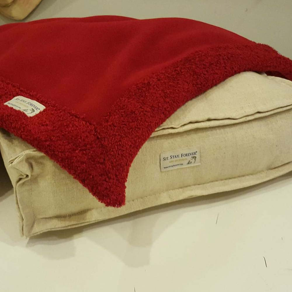 A folded Toxin Free Plush and Curly Velour Throw in red, placed on a beige cushion. Both the throw and the cushion have labels reading Sit.Stay.Forever.