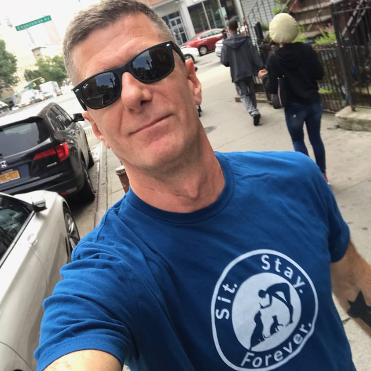 Man with sunglasses walking in the street wearing a blue colored 100% Cotton T shirt with Sit.Stay.Forever. logo in the center