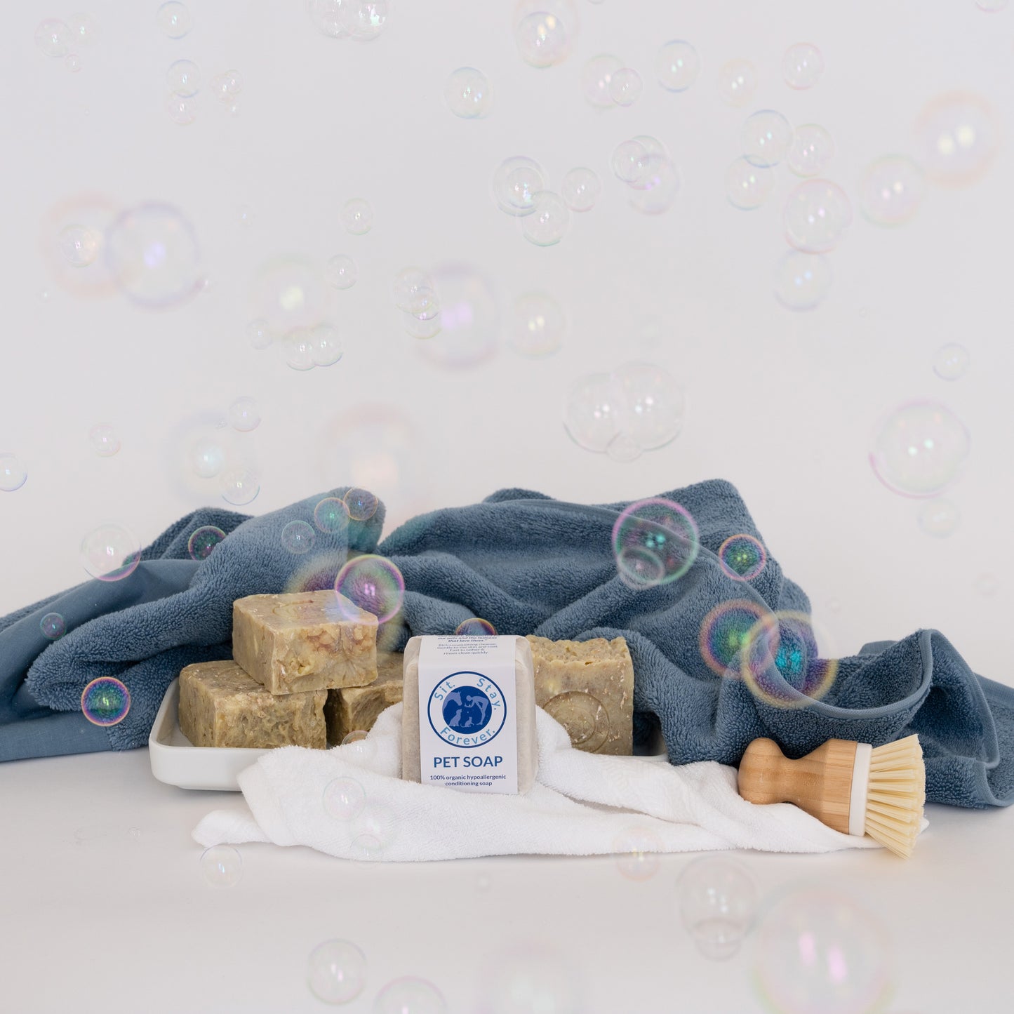 A tray with Sit.Stay.Forever pet soap bars, blue and white towels, and a wooden brush, surrounded by bubbles.