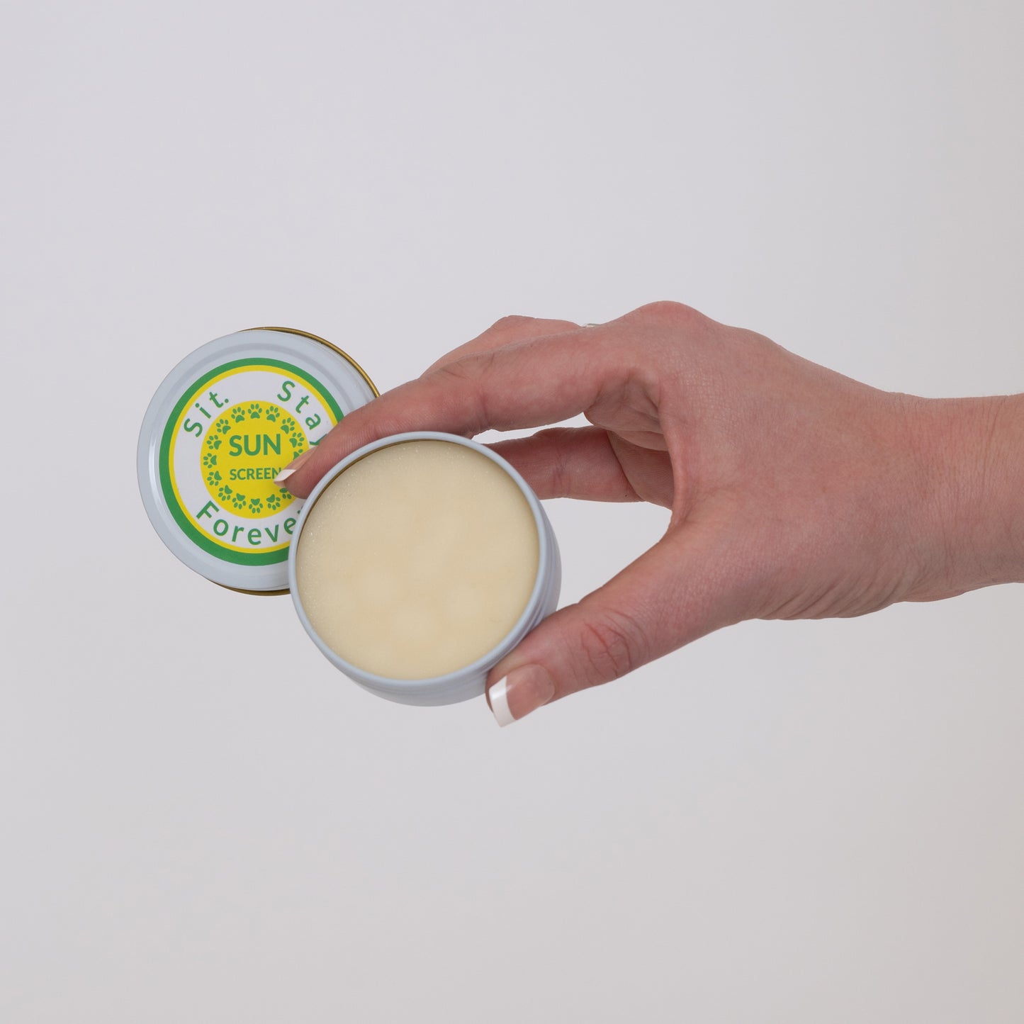 A hand holding an open tin of Sit.Stay.Forever sunscreen, showing the contents inside.